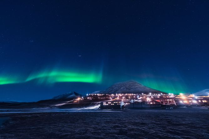 Northern lights illuminate the night sky over a mountain village, with numerous lights visible from buildings below.