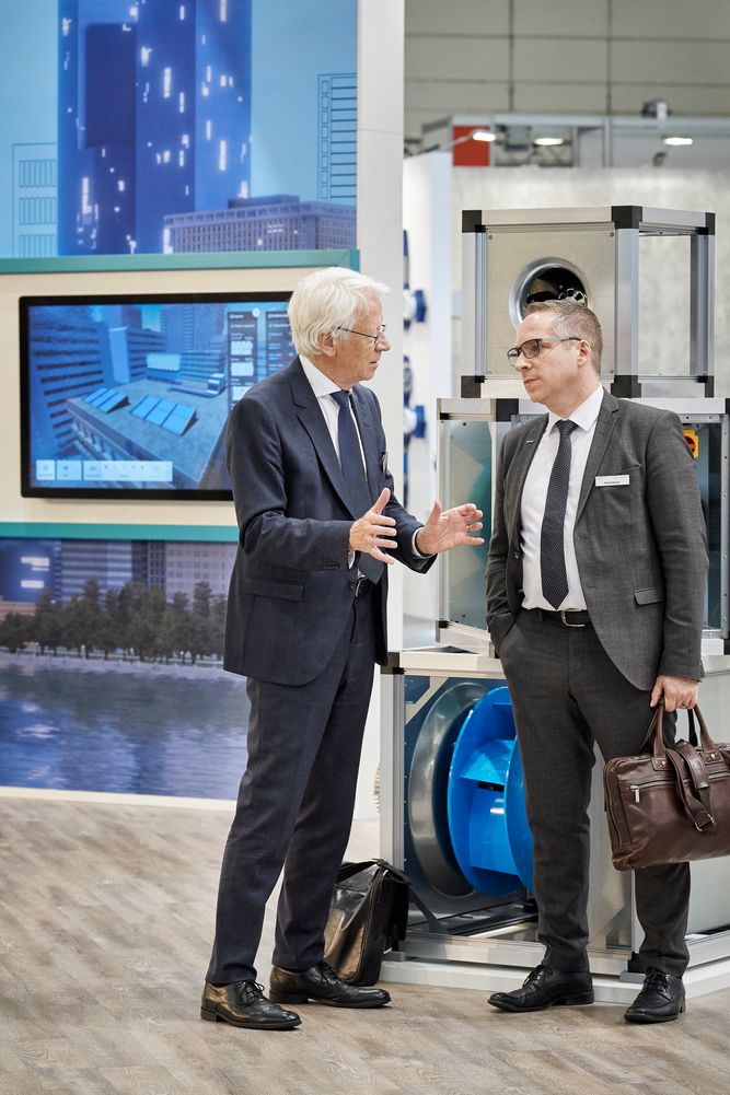 Gerald Engström and Roland Kasper are having a conversation at a trade show booth, with digital displays and industrial equipment in the background. One holds a briefcase.