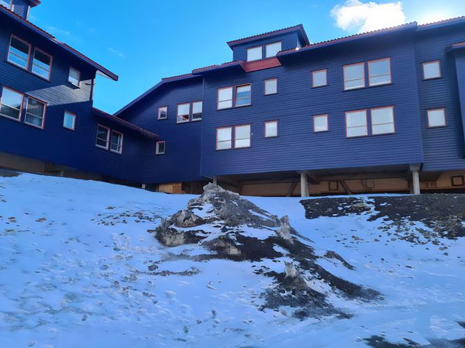 View of a blue multi-story building with numerous windows, situated on a snowy slope under a clear blue sky.