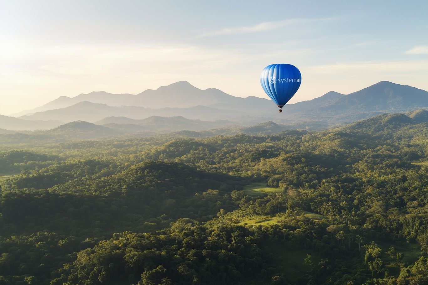 A blue hot air balloon floats above a lush green landscape with distant mountains under a clear sky.